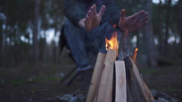 Guy sit on the chair, rubbing his hands to warm up near fire. Brutal unshaven man resting by the fire in the forest. — Stock Video