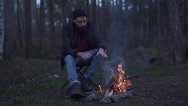 Homem barbudo sentar na floresta perto de fogueira e colocar bebida em uma xícara de garrafa térmica. Brutal unshaven cara descansando ao lado do fogo na floresta . — Vídeo de Stock