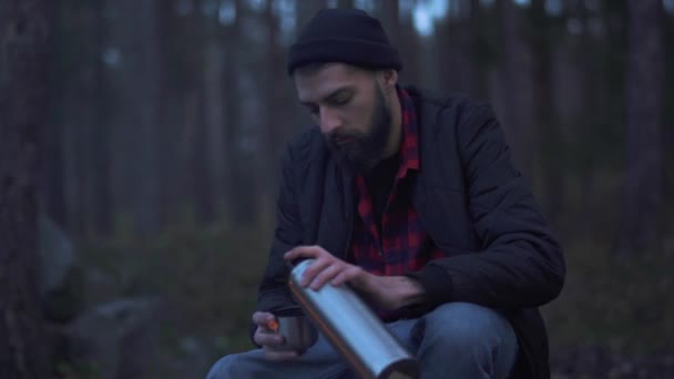 Young bearded man sit in the forest near bonfire and put beverage in a cup from thermos and drink. Brutal unshaven guy resting by the fire in the forest. — Stock Video