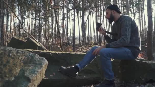 Бородатый парень точит топор в лесу. Жестокий человек сидит с топором на улице . — стоковое видео