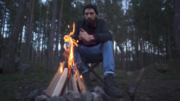 Un joven barbudo se sienta en la silla, frotándose las manos para calentarse cerca del fuego. Forestal junto al fuego en el bosque . — Vídeos de Stock