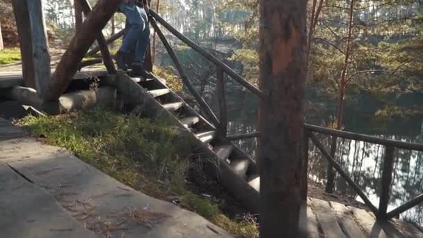 Hombre barbudo con hacha en las manos caminar por escaleras de madera cerca de bosque increíble con lago. Joven barbudo con un hacha al aire libre . — Vídeos de Stock