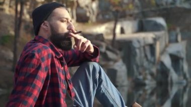 Ciddi sakallı adam oturup river ve yasaktır kahverengi puro yakınındaki ormanda. Acımasız Adam ekose gömlek ve mavi jeans açık havada bir puro sigara siyah sakallı.