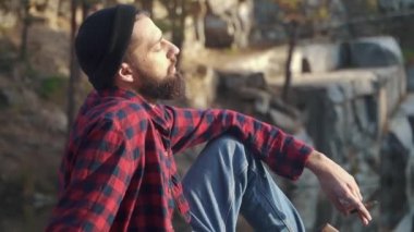 Sakallı adam oturup river ve yasaktır kahverengi puro yakınındaki ormanda. Ekose gömlek ve mavi jeans açık havada bir puro sigara siyah sakallı acımasız adam.