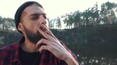 Genç adam oturup river ve yasaktır kahverengi puro yakınındaki ormanda sakallı.