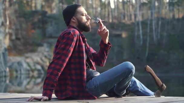 Um tipo barbudo sentou-se na floresta perto do rio e fumava charuto castanho. Brutal homem sério com uma barba preta em uma camisa xadrez e jeans azul fumar um charuto ao ar livre . — Vídeo de Stock