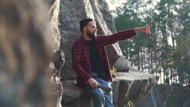 Young man with ax in hand sitting on the huge mountain snone on the edge of a cliff and pointing his finger. Brutal serious guy with ax outdoors. — Stock Video