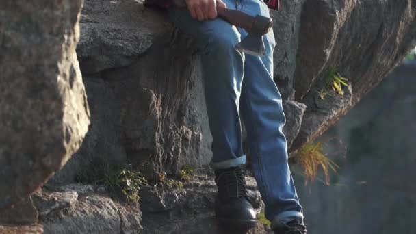 Bearded guy with axe sit near huge stone and looking into the distance. Brutal serious man with ax outdoors. — Stock Video