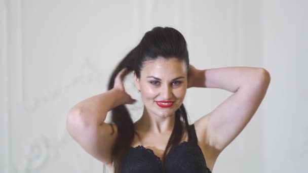 Hermosa chica sonriente en sujetador negro posando sobre un fondo blanco en el estudio. Linda chica en lencería posando en un estudio brillante. Mujer encantadora sonríe y endereza el cabello . — Vídeo de stock