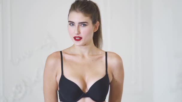 Tall slim girl in a dark bra posing on a white background in a bright studio. Cute girl smiles and sends air kiss. Cute girl flirting and sends air kiss. — Stock Video