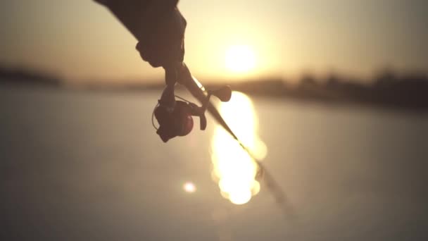 Mão masculina segura vara de pesca no fundo do rio durante o pôr do sol close-up. Pescador segura uma vara de pesca contra o pôr do sol. Luz solar através de uma vara de pesca. Pesca fluvial . — Vídeo de Stock