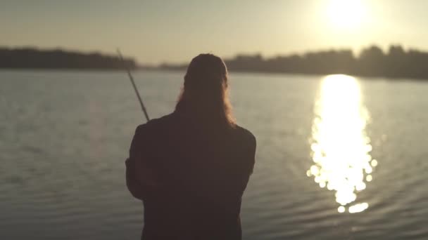 Ψαράς Ψάρεμα με καλάμι στο φόντο του ποταμού κατά το ηλιοβασίλεμα. Fisher ενεργοποιεί το πηνίο ένα περιστρεφόμενο τραβώντας έξω τα ψάρια. Ψάρεμα σε ποτάμι. — Αρχείο Βίντεο