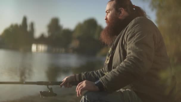 Bärtige Fischer, die am Ufer des Flusses fischen. Ein Mann sitzt mit einer Angelrute im Freien. Flussfischerei. — Stockvideo