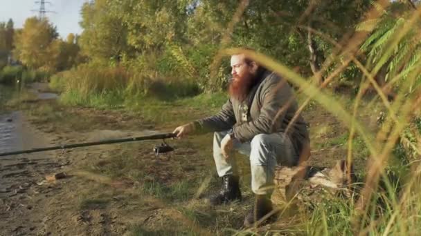 Visser zit op een logboek op de oever van de rivier, het roken van een sigaret, holding van een hengel in zijn hand en zijn horloge kijken. Riviervissen. — Stockvideo