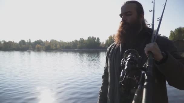 Pescador barbudo con largas barbas pasea por la orilla del río con cañas de pescar. Movimiento lento . — Vídeo de stock