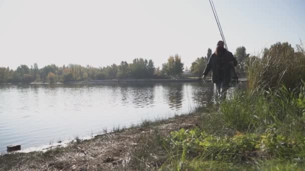 Pêcheur solitaire à la barbe longue marche sur la rive de la rivière avec des cannes à pêche. L'homme regarde au loin, couvrant ses yeux avec sa main du soleil. Mouvement lent . — Video