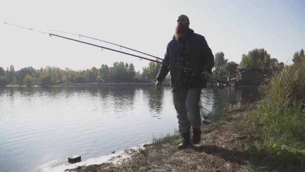 Pescador adulto con largas barbas pasea cerca del río con cañas de pescar y red de pesca. El hombre mira a lo lejos, cubriéndose los ojos con la mano del sol — Vídeo de stock