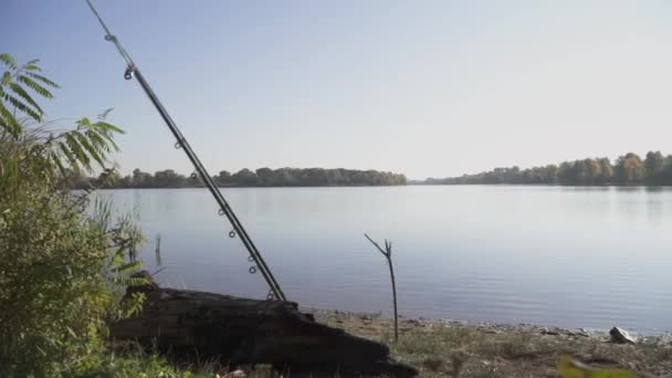 在平静的水面、绿树和干净的蓝天的背景下, 两根钓鱼竿躺在河岸上. — 图库视频影像