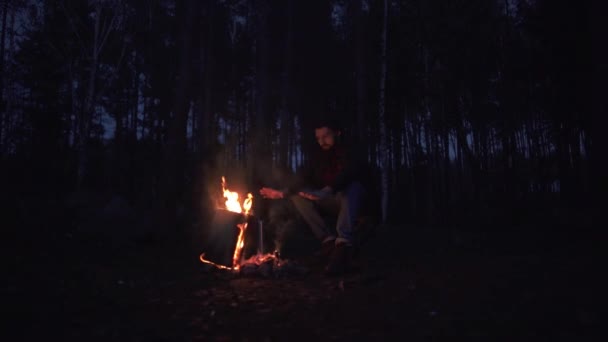 Un joven barbudo se sienta en una silla y se calienta las manos junto al fuego. Forestal junto al fuego en el bosque. Brutal chico por hoguera al aire libre . — Vídeo de stock