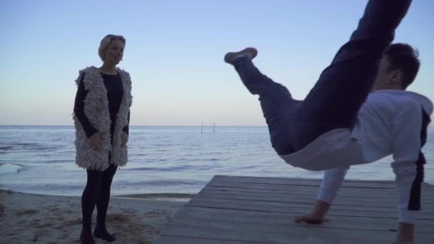 Aziatisch vent in casual kleding dansen breakdance voor zijn mooie Kaukasische vriendin in de buurt van het meer. Schattige lachende meisje kijkt naar haar geliefde met bewondering en liefde. Slow motion. — Stockvideo