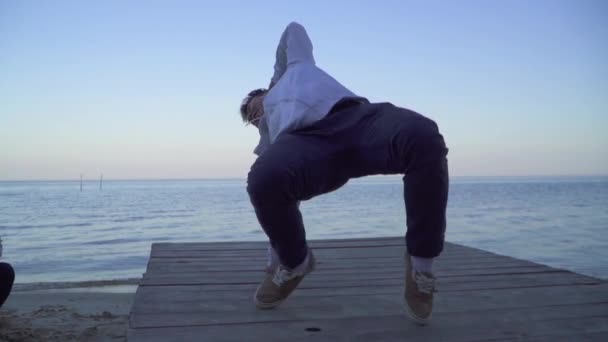 Guapo asiático chico en ropa casual bailando break dance delante de su hermosa novia caucásica en la orilla del río. Hombre asiático bailando breakdance al aire libre. Linda chica sonriente mira a su amante — Vídeos de Stock