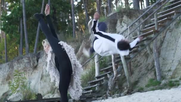 Asiatisk kille och kaukasiska flicka dansa modern dans. Tonåringar i casual kläder dans på den sandiga stranden av floden nära trätrappor. Aktiva interracial par dansar i fri stil — Stockvideo