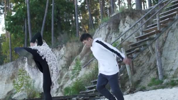 Teenageři v ležérní oblečení, tanec na písečné břehu řeky poblíž dřevěné schody. Asijské chlap a kavkazské dívka tančí současného tance. Aktivní pár interracial tančí ve volném stylu — Stock video