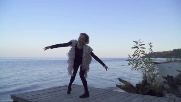 Красива молода біла жінка в повсякденному одязі танцює сучасний танець на відкритому повітрі. Гнучка дівчина в щільних легінсах танцює на дерев'яній платформі на фоні поверхні води . — стокове відео