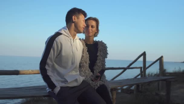 Interracial young couple in love makes selfie sitting on a wooden bench in the sunshine on the background of a lake. Asian guy kisses his caucasian girlfriend on a date outdoors. — Stock Video