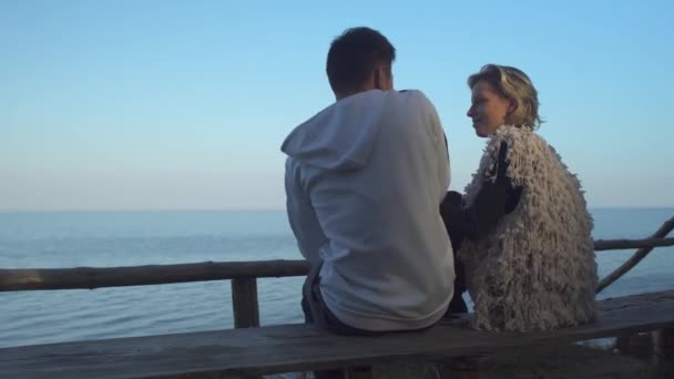 Young guy and girl are sitting on a bench and hugging on the background of the water surface. Young couple in love on a date outdoors. Relationship of a young guy and a girl. — Stock Video