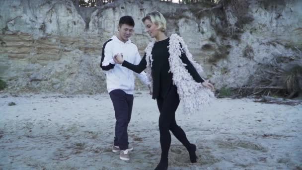 Interracial par Dans i fri stil utomhus. Asiatisk kille och kaukasiska flicka dansa modern dans. Tonåringar i fritidskläder som dansar på den sandiga stranden av floden. Slow motion. — Stockvideo