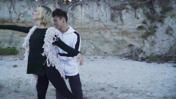 Pareja de jóvenes bailarines bailando en estilo libre al aire libre. Hombre asiático y chica caucásica bailando danza contemporánea. Adolescentes en ropa casual bailando en la orilla arenosa del río cerca de las escaleras de madera — Vídeos de Stock