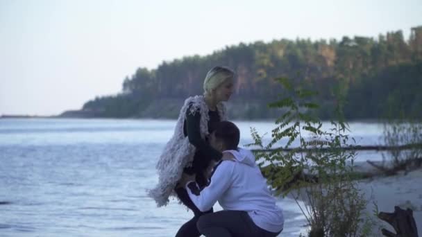 Asiatische Kerl hebt seine kaukasische Freundin über seinen Kopf auf dem Hintergrund eines Sees und Waldes. Jugendliche in Freizeitkleidung stehen am sandigen Flussufer. Exotisches Paar im Freien. — Stockvideo