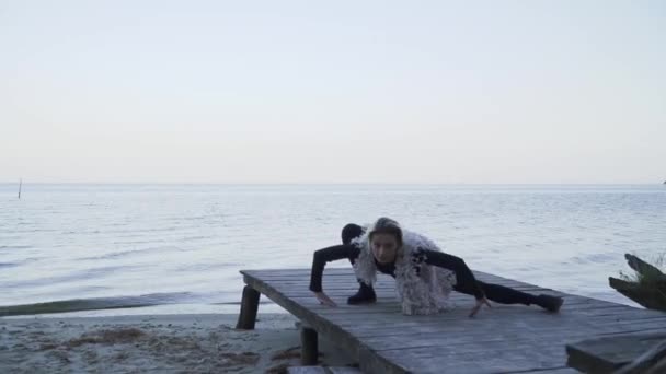 Молода біла жінка в повсякденному одязі танцює сучасний танець на відкритому повітрі. Гнучка дівчина в щільних легінсах танцює на дерев'яній платформі на фоні поверхні води . — стокове відео