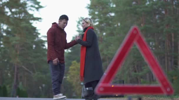 Um casal a jurar no fundo de um carro avariado. Triângulo de aviso em primeiro plano do cara e da menina jurando . — Vídeo de Stock