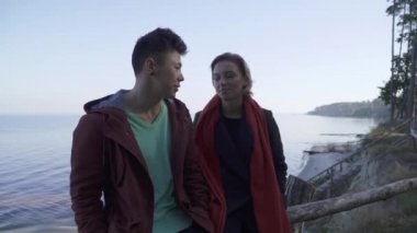 Interracial kaç rahat içinde gülümseyen genç çam ağaçları ve bir göl bir arka plan üzerinde uçurumun başına oturmuş giymek. Çok etnik çift bir tarih, mutlu bir şekilde gülümseyerek ve konuşurken zevk aşık