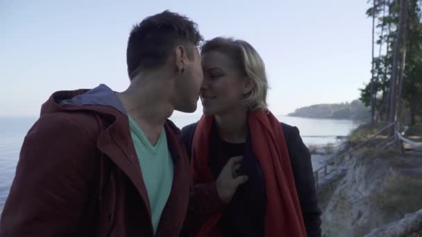 Bella giovane coppia multietnica sorridente innamorata che si bacia su uno sfondo di una scogliera, pini e un lago. baci interrazziale coppia all'aperto . — Video Stock