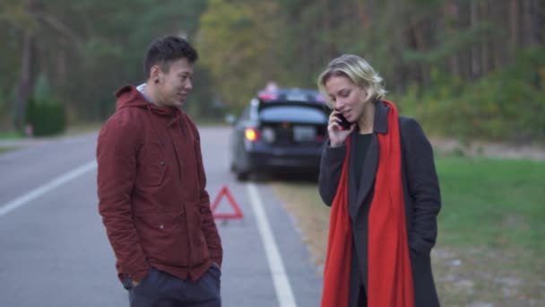 Un jeune couple debout sur le fond d'une voiture garée sur le bord de la route. Belle fille aux cheveux blonds parle sur le téléphone portable tout en se tenant près de son mari à l'extérieur. Relation entre : — Video