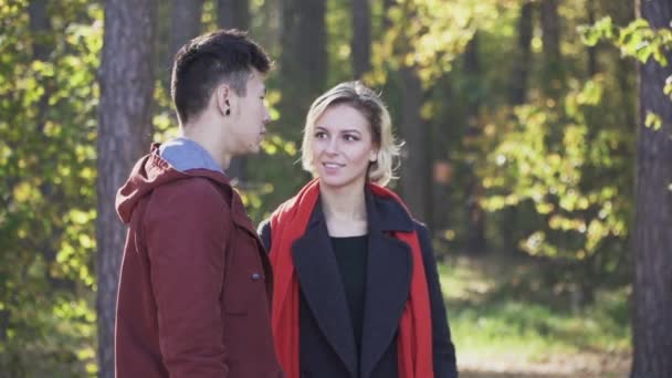 Une belle jeune fille ludique aux cheveux blonds dans un manteau et une écharpe rouge s'enfuit de son petit ami dans la forêt. Le gars court après la fille à travers la forêt de pins au soleil. Loisirs d'un jeune — Video