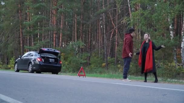 Multiethnisches Paar auf dem Hintergrund eines kaputten Fahrzeugs, das am Straßenrand steht. schöner asiatischer Mann, der den Daumen nach oben hält, um ein Auto zu erwischen. Mann versucht Auto auf Straße zu stoppen. — Stockvideo