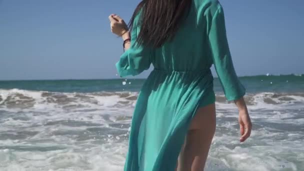 Mujer linda con un cuerpo perfecto vestido de turquesa disfruta de las olas del mar de pie en el agua. La ola rueda sobre una chica en un vestido de verano ligero y le da mucho placer . — Vídeos de Stock