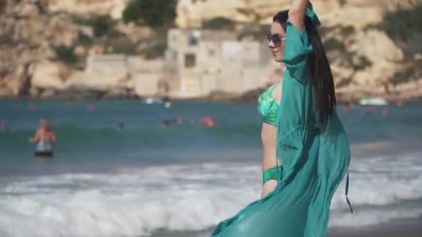 Hermosa chica de cabello oscuro y cuerpo perfecto con gafas de sol, vestido transparente y bikini disfrutando de sus vacaciones sonriendo y caminando por la playa del mar en un día soleado de verano. Joven chica feliz — Vídeo de stock