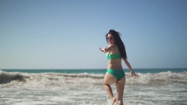 Beautiful girl with a perfect body wearing bikini walks into the sea and turns to the camera with inviting gesture. Sweet girl invites to follow her. — Stock Video