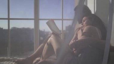 Genç çıplak sarışın adam dan kat tavan tül ile birlikte pencere yakınındaki kitap okurken sarılma güzel seksi çıplak kadın. Genç ve seksi çift aşık.