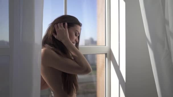 Jeune belle brune nue avec de longs cheveux debout près de la fenêtre à l'intérieur de la maison. Sensuelle et passionnée. Beauté femme avec corps attrayant en culotte noire . — Video