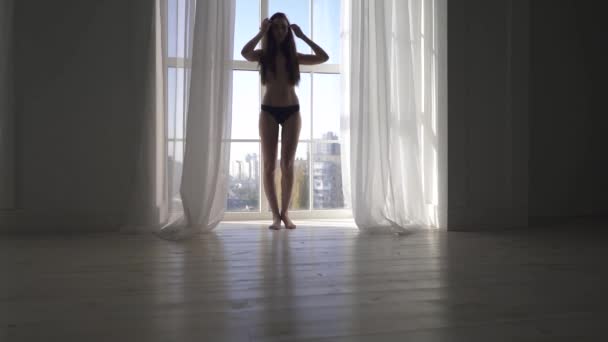 若いスリム 床から天井までの大きな窓の近くの長い茶色の髪のスタンドと裸のセクシーな女の子です 女性のストレッチ — ストック動画