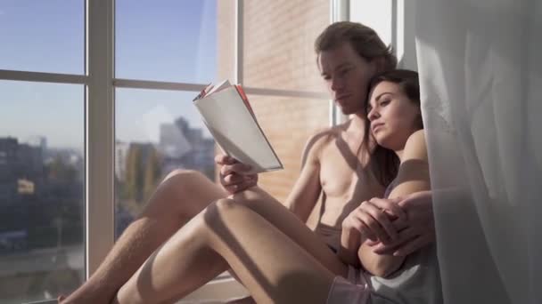 Beautiful sexy naked woman hugging young nude blond man while reading a book together near window from floor-to-ceiling through tulle. Young and sexy couple in love embrace. — Stock Video