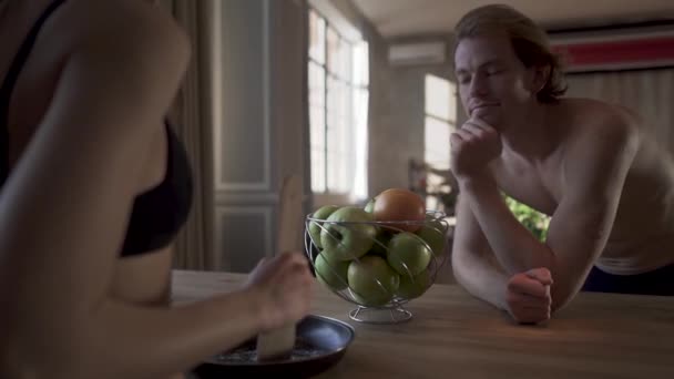 Giovane coppia nuda stare vicino al tavolo da cucina, ragazza sexy in lingerie mescolare il sale in padella e battere con cucchiaio il suo uomo in biancheria intima. Uomo sorriso indietro . — Video Stock