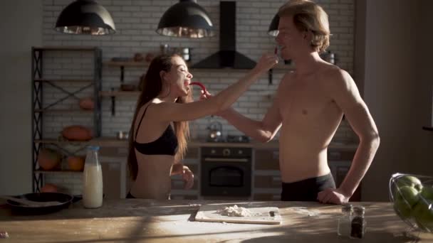 Joven chica caliente sexy en sujetador y hombre guapo en la cocina se alimentan entre sí picante chile rojo pimienta. Amar pareja en ropa interior divertirse en la cocina. Movimiento lento . — Vídeos de Stock