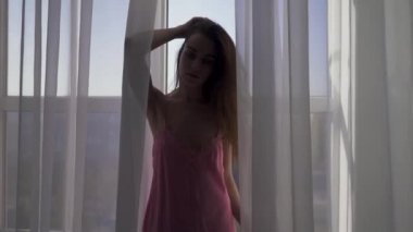 Tatlı genç şehvetli kadın geceliği büyük beyaz pencere yanında poz uzun kahverengi saçlı. Kusursuz bir beden pijama ile kız.